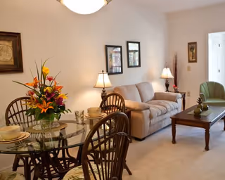 A cozy living room and dining area featuring a beige sofa, a wooden coffee table with decorative items, a green armchair, and a glass-top dining table with four wooden chairs. The dining table has a colorful floral centerpiece and place settings. The walls are decorated with framed artwork and two table lamps provide warm lighting.