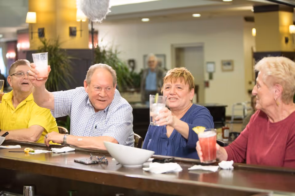 A group of older adults sitting at a bar counter in a senior living facility raising their drinks in a toast.
