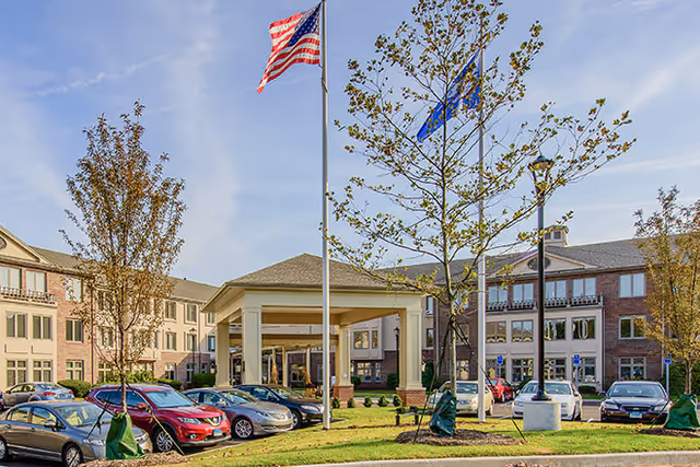Exterior view of Brookdale Chatfield senior living facility showing a multi-story brick building with numerous windows, a covered entrance with columns, several parked cars in the parking lot, and three flagpoles with the American flag and other flags flying. Trees and landscaping are visible around the parking area under a clear sky.
