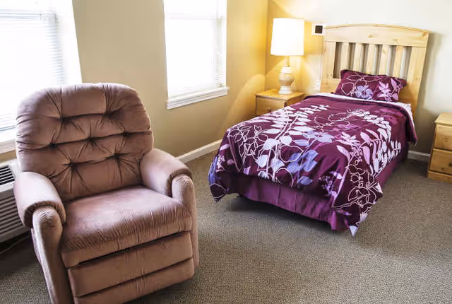 A cozy senior living bedroom featuring a single wooden bed with a purple floral bedspread, a matching pillow, a wooden nightstand with a lamp, a brown cushioned recliner chair, and two windows letting in natural light.