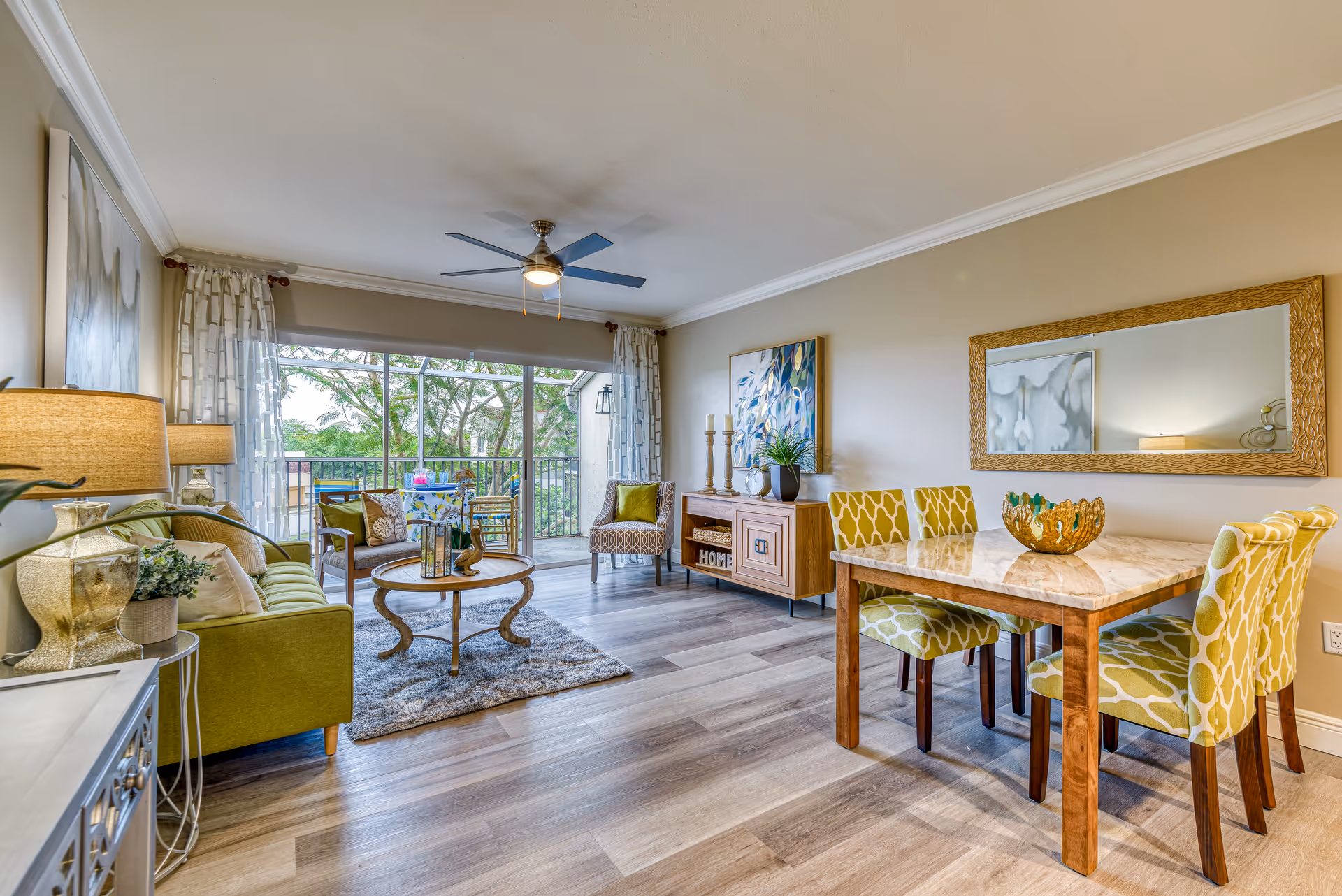 Bright open living and dining area with a green sofa, round coffee table, wooden dining table with patterned chairs, and sliding glass doors to a balcony.