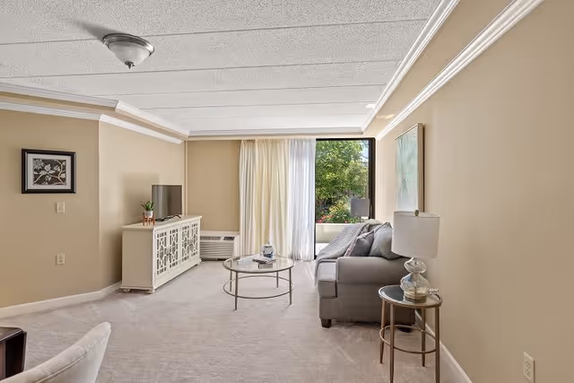 A bright living room with beige walls and carpet, featuring a gray sofa with cushions, a round glass coffee table, a side table with a lamp, a white TV stand with a flat-screen TV, and a large window with sheer curtains letting in natural light and showing greenery outside.
