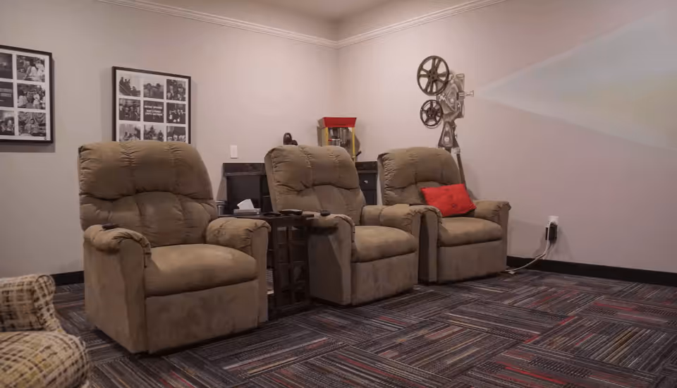 A cozy room with three beige recliner chairs arranged in a row facing a wall where a vintage film projector is projecting light. The room has patterned carpet flooring, framed black and white photos on the wall, and a small table with a popcorn machine behind the chairs.
