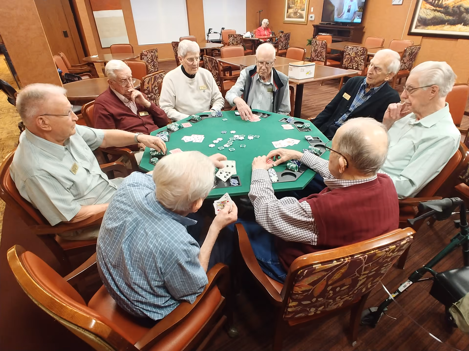 A group of elderly men sitting around a green poker table playing cards and poker chips in a well-lit room with wooden floors and patterned chairs. In the background, there is a woman sitting at another table and a television mounted on the wall.