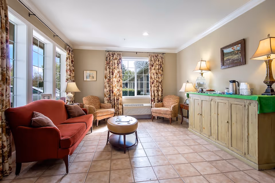 A cozy living room with a red sofa, two patterned armchairs, a round ottoman, and a long wooden cabinet topped with lamps, a water dispenser, and coffee supplies. The room has large windows with floral curtains letting in natural light, beige walls, and tiled flooring.