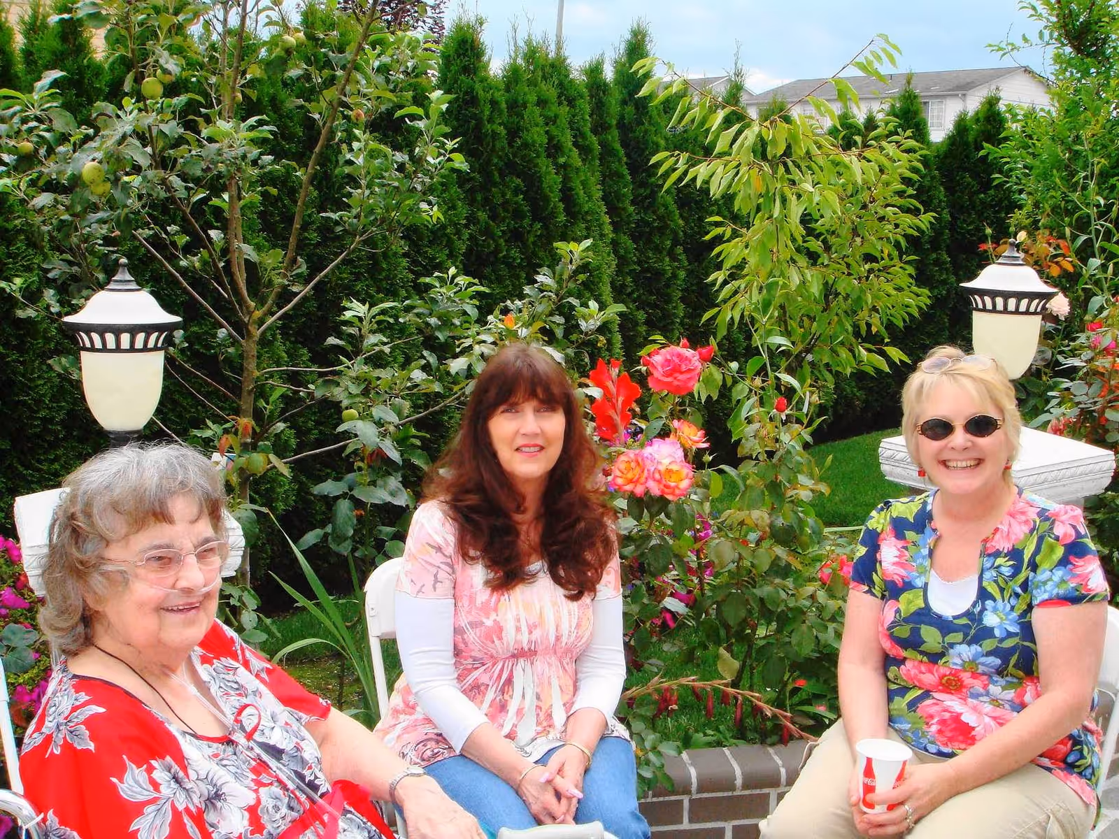 Three women sitting outdoors in a garden area with lush green trees and colorful flowers around them. They are smiling and appear to be enjoying a pleasant day. Two white garden lamps are visible in the background.
