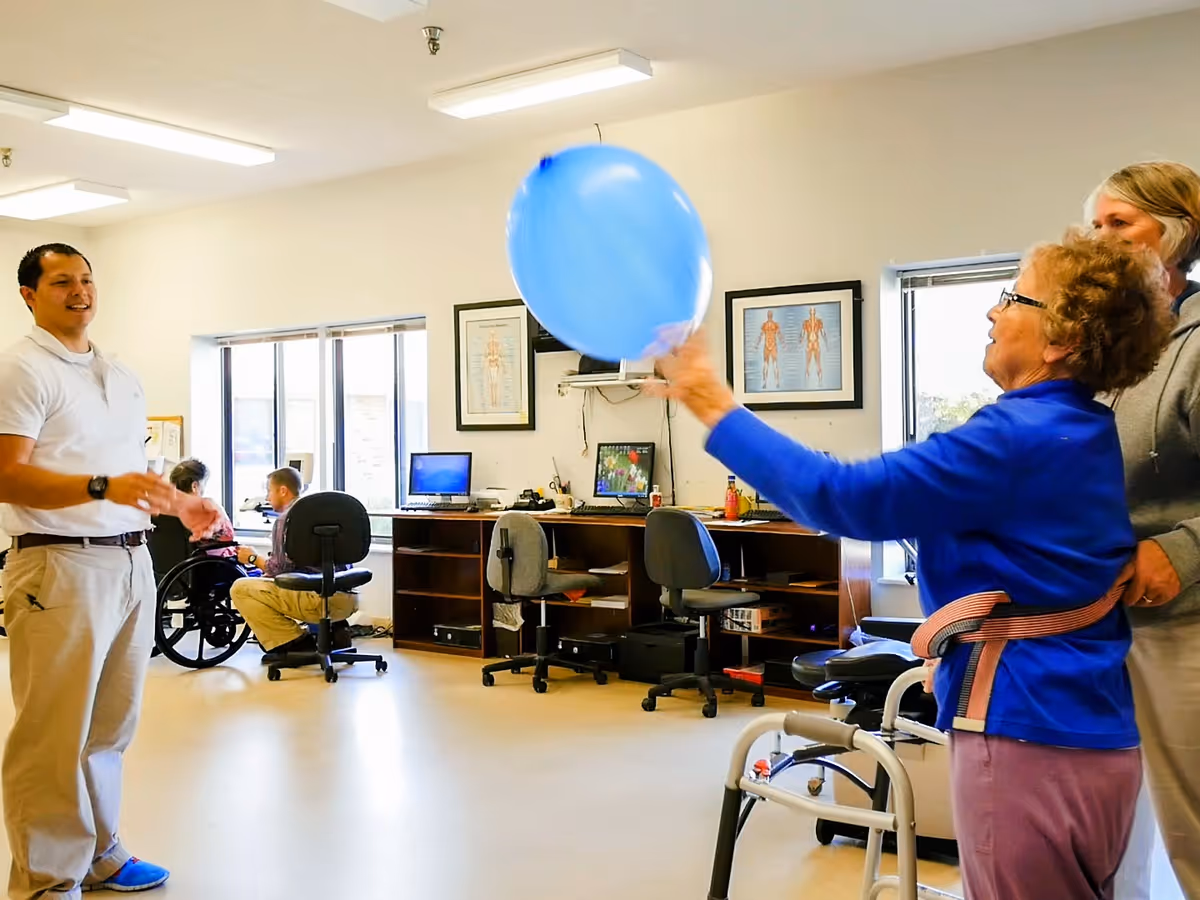 An elderly woman using a walker and wearing a support belt plays with a large blue balloon indoors, assisted by a caregiver. A man in a white shirt and khaki pants stands nearby, smiling. In the background, two other people are seated, one in a wheelchair and another on an office chair, in a room with desks, computers, and anatomical charts on the wall.