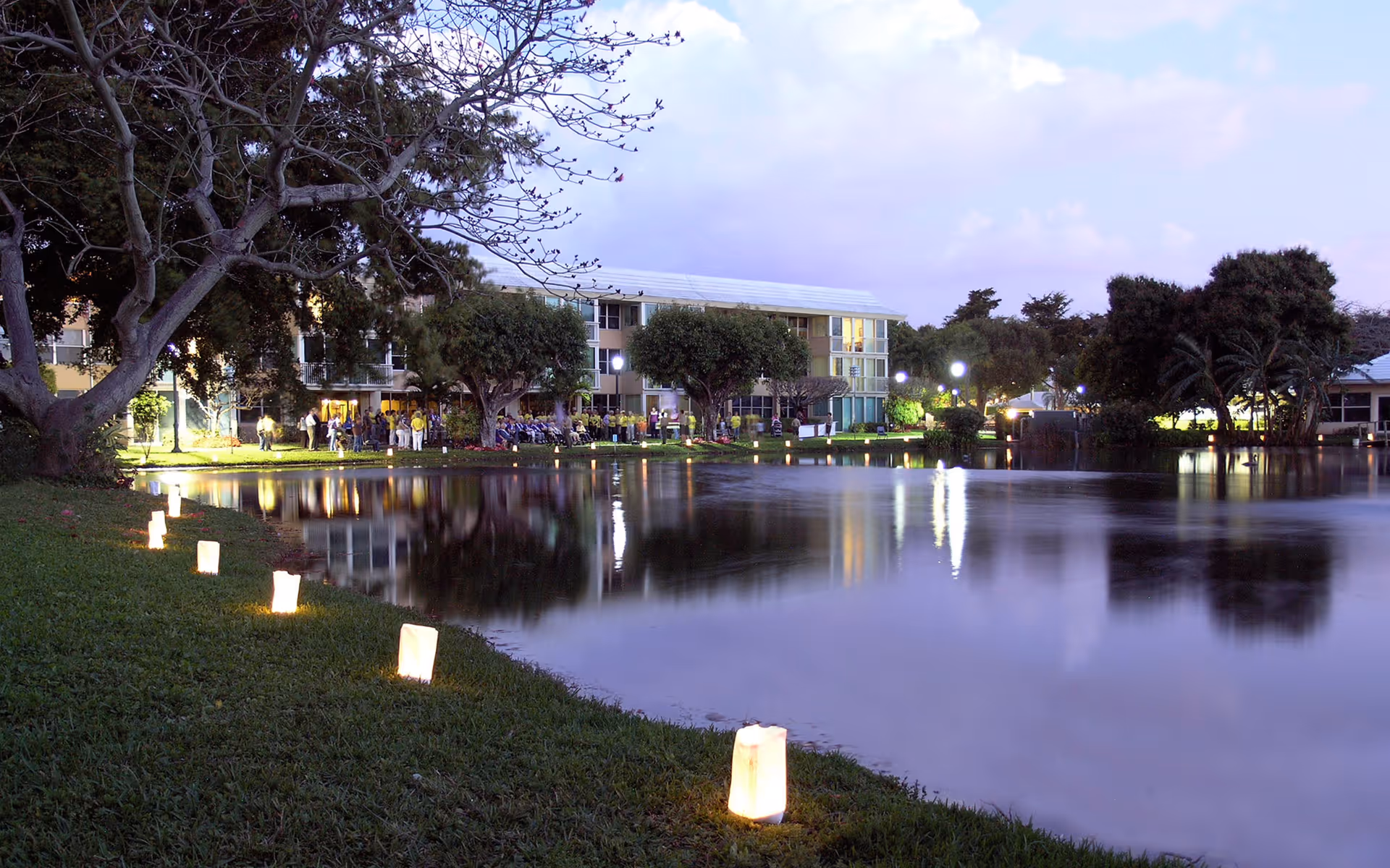 Evening view of a calm pond with illuminated lanterns lining the grassy shore. In the background, a multi-story building is visible with trees and outdoor lighting, reflecting on the water's surface.