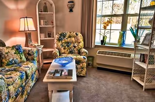 A cozy living room with floral patterned sofa and armchair, a white coffee table with magazines and a decorative blue bowl, a white bookshelf with various items, a table lamp, and a window with beige curtains letting in natural light. There is also a white wicker shelving unit and a heating/cooling unit beneath the window.