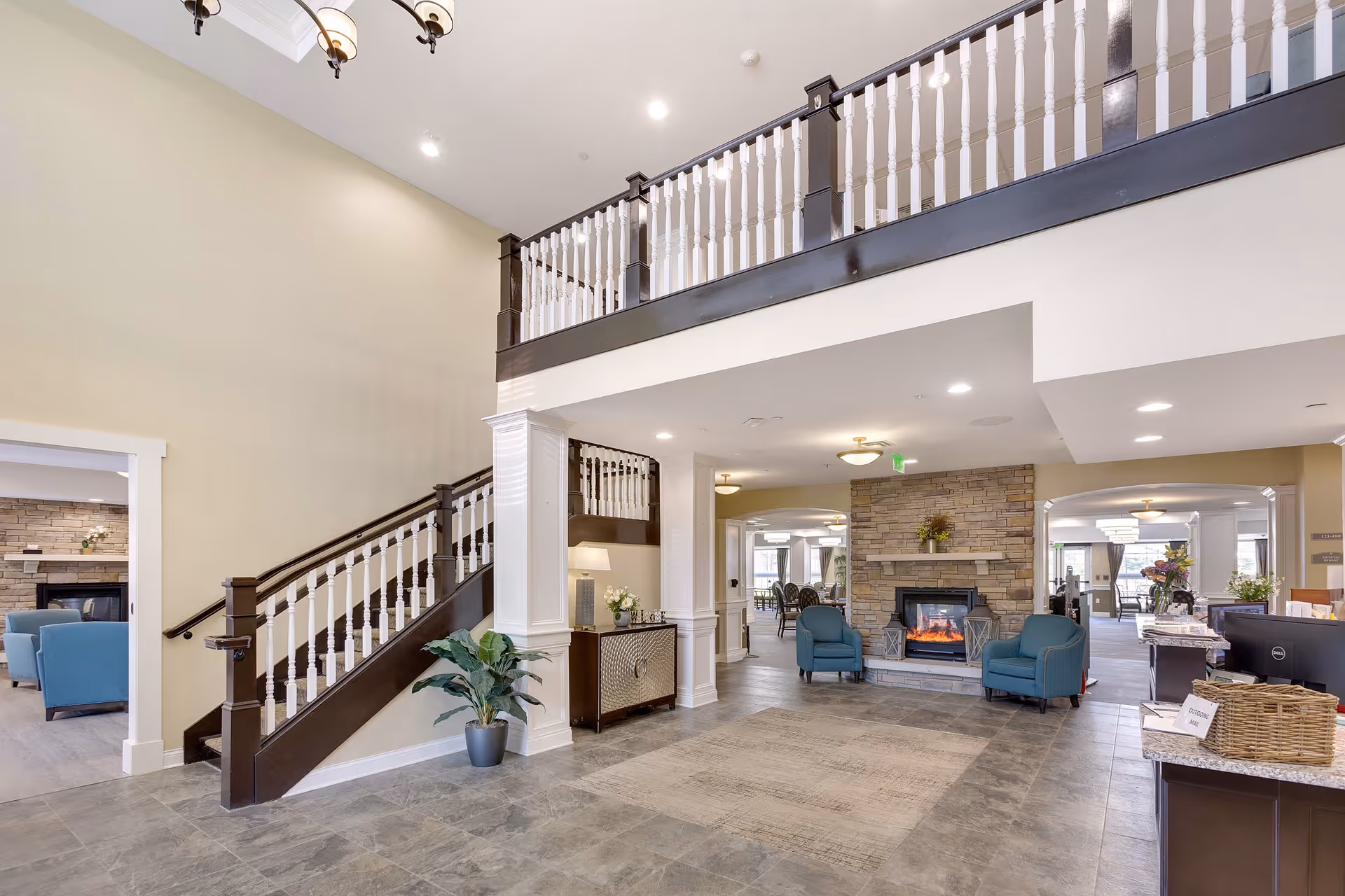 Spacious and well-lit interior of a senior living facility featuring a stone fireplace with two blue armchairs in front, a staircase with dark wood and white railings leading to an upper level, and a reception desk on the right side. The area has tiled flooring, beige walls, and decorative plants, with adjoining rooms visible in the background.