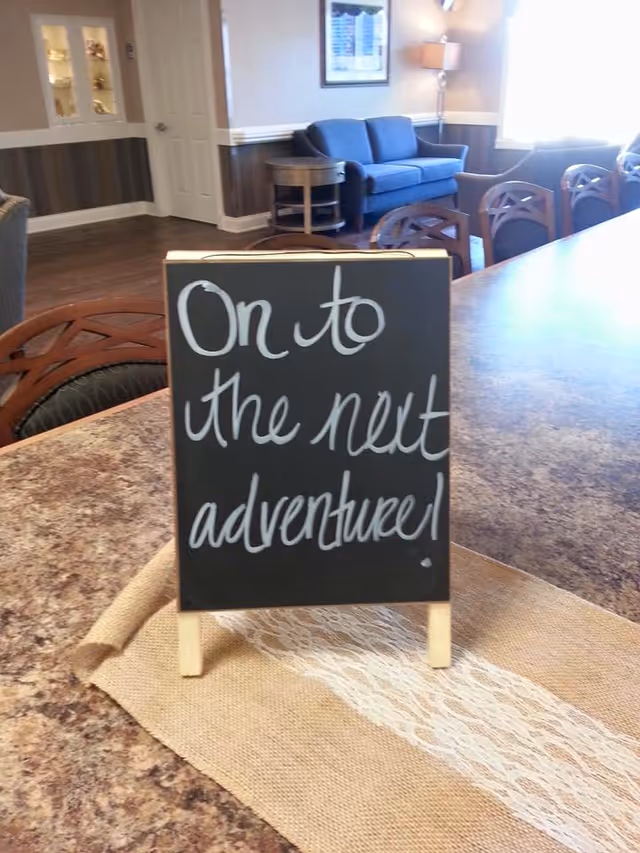 Small chalkboard on a countertop reading "On to the next adventure!" in a communal living area with chairs and a sofa in the background.