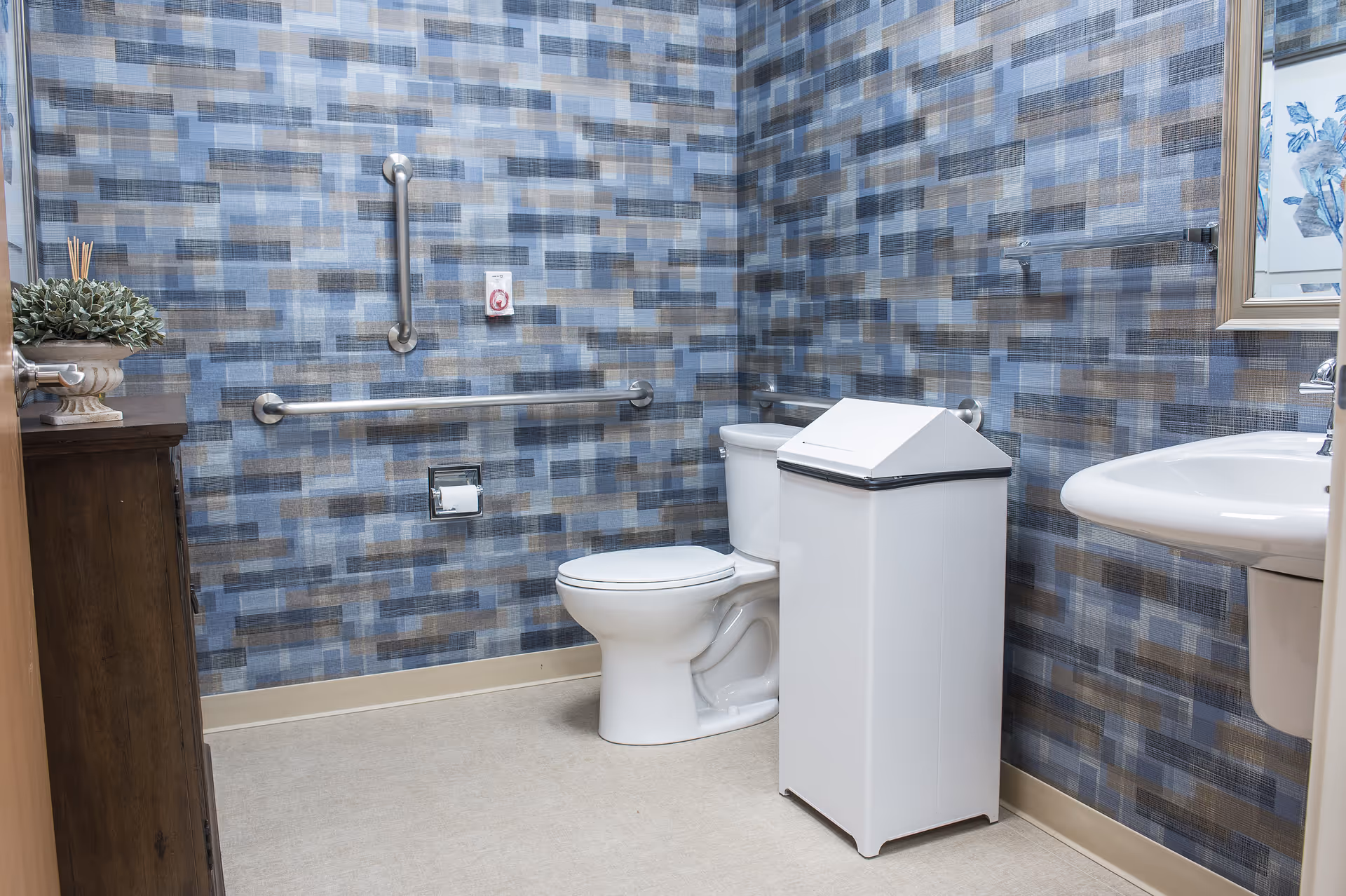 A clean bathroom with a white toilet, white trash bin, and a white sink. The walls are covered with a blue, gray, and beige patterned wallpaper. There are metal grab bars on the walls for support, a wooden cabinet with a potted plant on top, and a framed floral picture hanging on the wall.