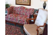 A cozy living room area featuring a floral patterned sofa, a cushioned armchair with wooden arms, a wooden side table with a lamp, and a framed picture on the wall.