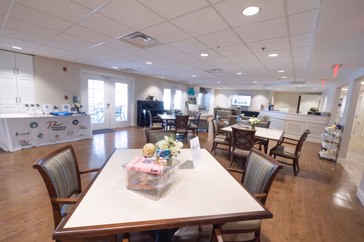 A spacious and well-lit common area in a retirement community featuring multiple tables and chairs arranged for socializing or activities. The room has wooden flooring, a piano against the wall, a table with promotional materials, and a television mounted on the far wall. Large windows and glass doors allow natural light to enter the room.