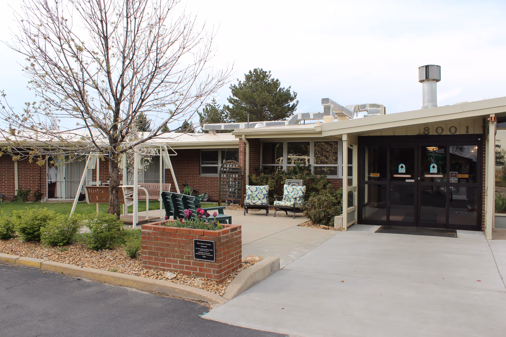 Exterior view of Arbor View Skilled Nursing Division showing the entrance with glass doors, a small garden area with a tree, benches, and chairs outside the building under a cloudy sky.