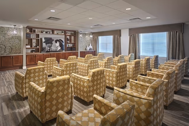 A cozy media room with multiple rows of patterned armchairs facing a large flat-screen TV mounted on a wooden entertainment center. The room has two windows with curtains, soft lighting, and a carpeted floor.