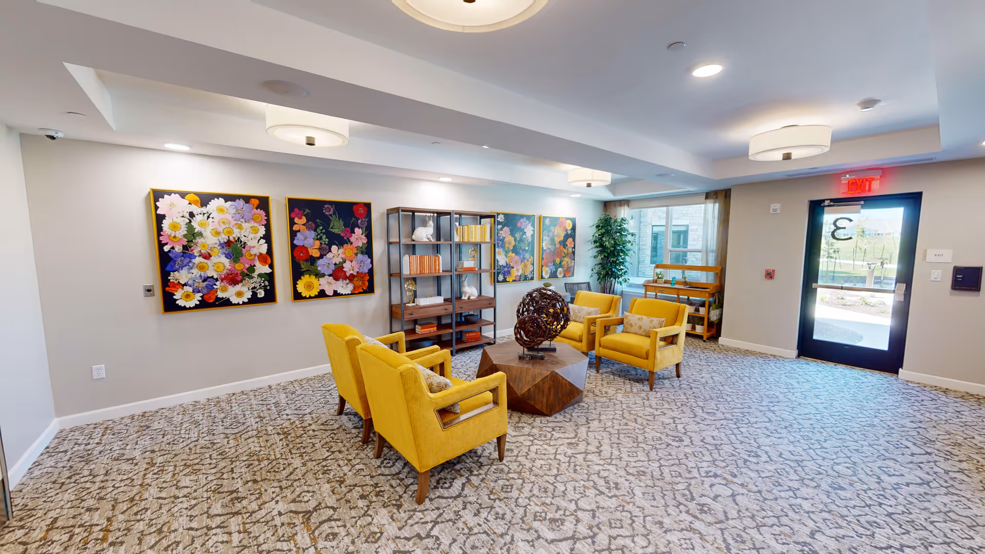 Bright senior living lounge with yellow armchairs around a wooden coffee table, floral artwork on the wall, and an exit door to the right.