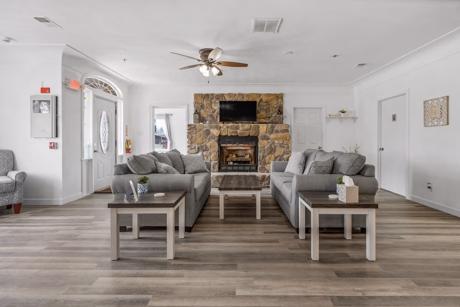 Bright communal living room with two gray sofas facing a stone fireplace and wall-mounted TV, coffee tables, and wood-look flooring.