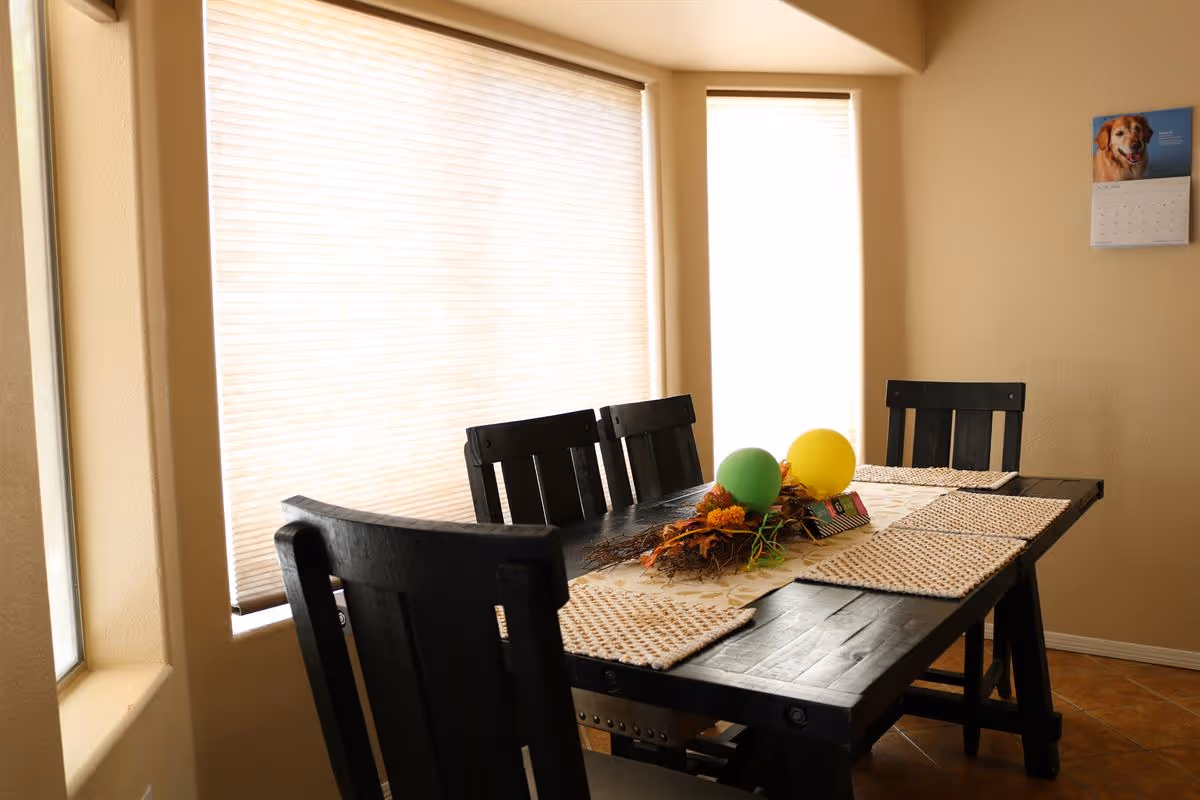 A dining room with a dark wooden table and four matching chairs. The table is decorated with a centerpiece featuring two balloons, one green and one yellow, along with some autumn-themed foliage. The room has beige walls and two large windows with closed blinds, allowing soft natural light to fill the space. A calendar with a dog picture is hanging on the wall.