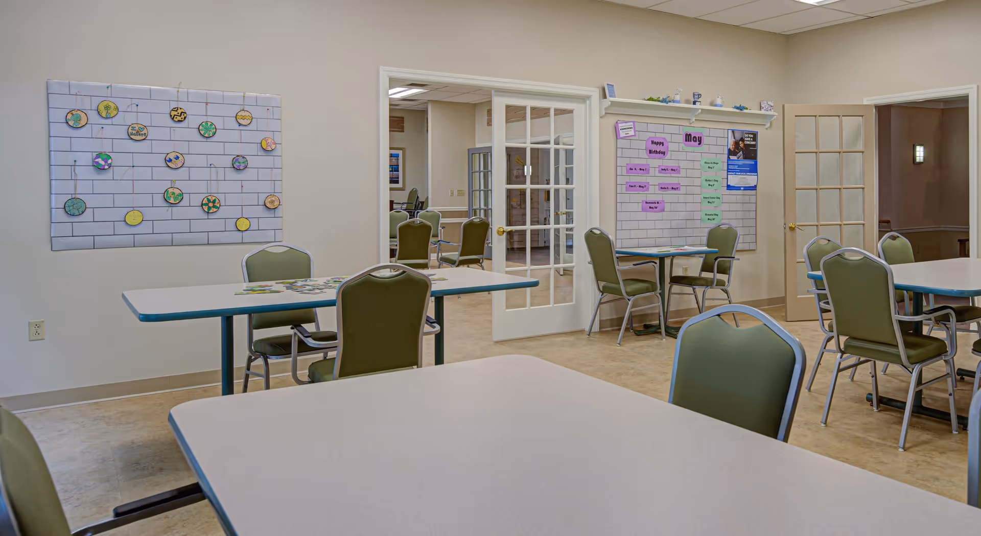 A well-lit room with several tables and green chairs arranged for group seating. The walls have bulletin boards with colorful decorations and notes, including a calendar for May and a birthday reminder. There are glass-paneled double doors leading to another room with more chairs and tables visible.