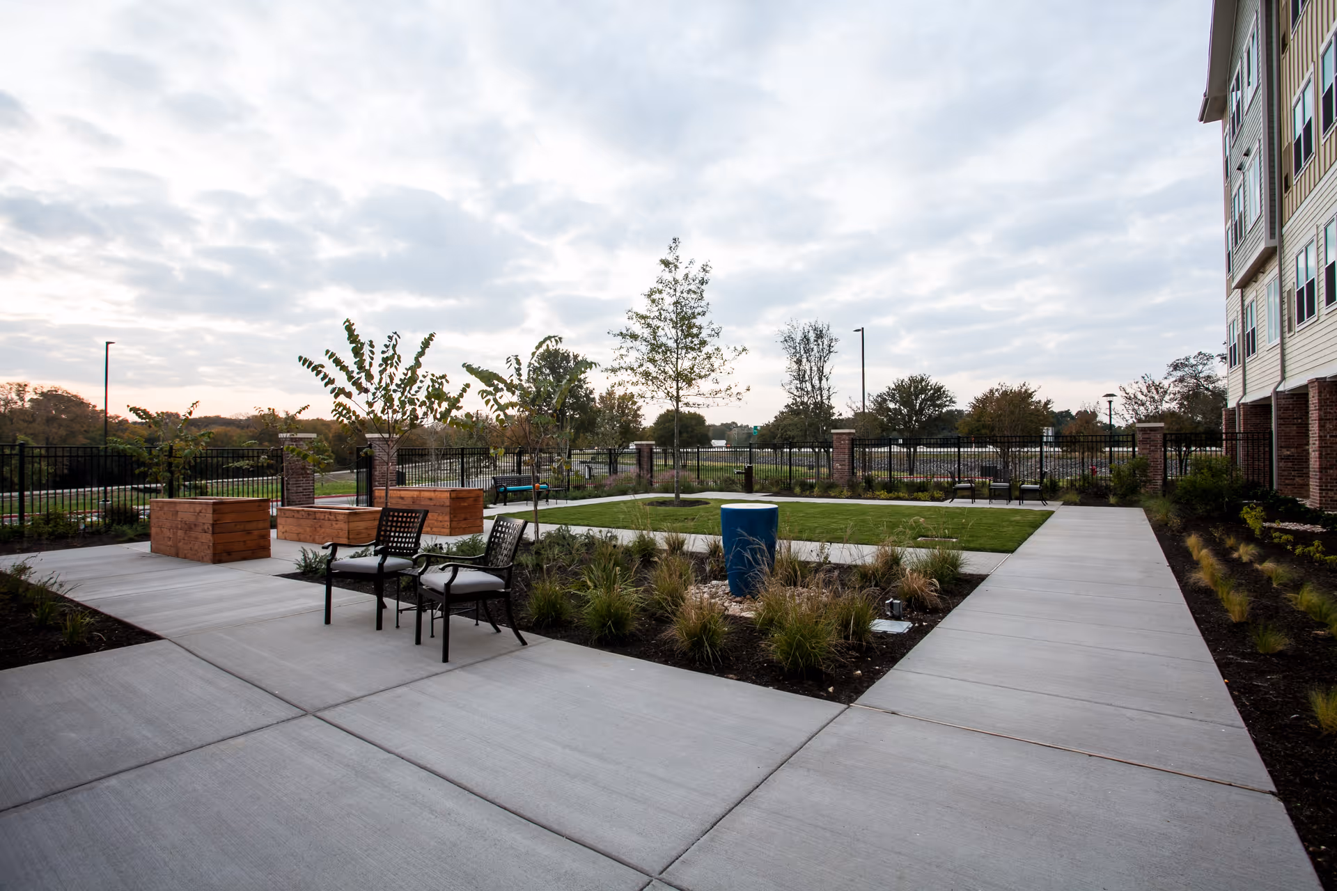 Outdoor patio area at The Delaney at Lake Waco featuring concrete walkways, metal chairs with cushions, wooden planter boxes, small trees, and landscaped grass and plants, with a partly cloudy sky overhead.