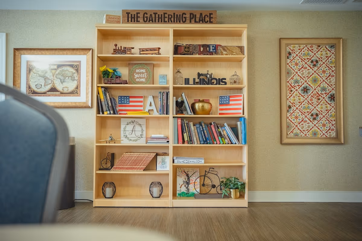 A wooden bookshelf labeled The Gathering Place filled with books, American flags, and decorative items between framed wall art in a seating area.