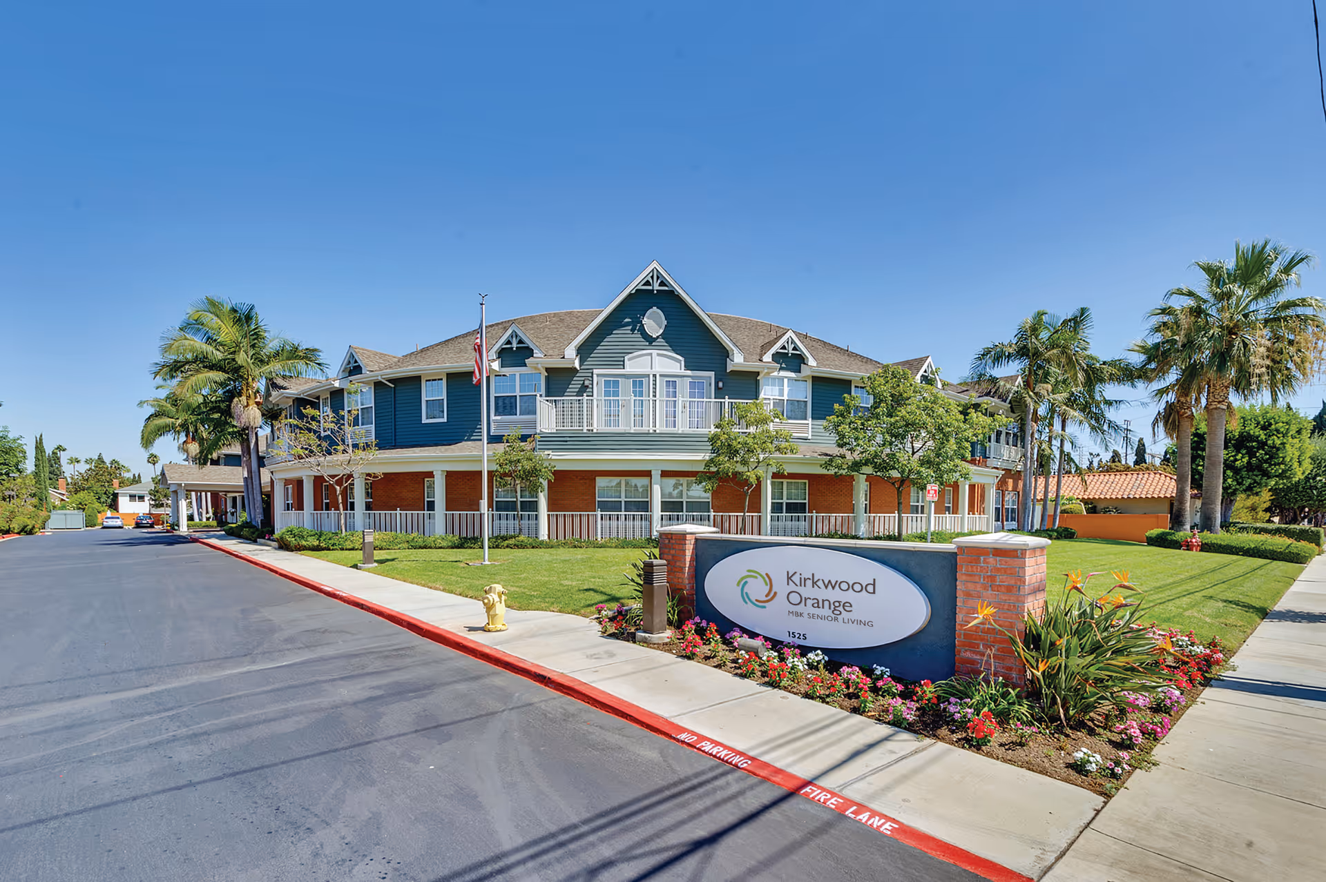 Front exterior of a two-story senior living building with palm trees, a lawn, and a 'Kirkwood Orange' sign.