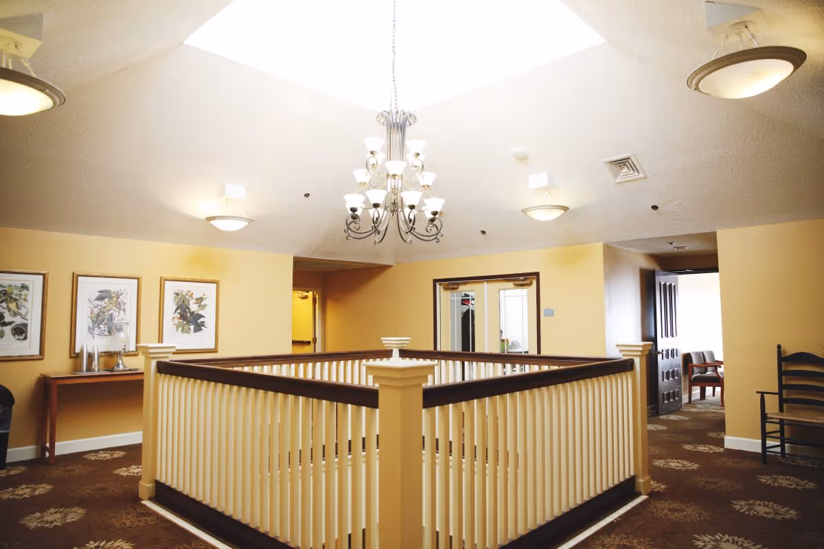 Interior view of a senior living facility hallway with a central staircase surrounded by a white and dark wood railing. The ceiling features a chandelier and recessed lighting. The walls are painted yellow, and there are framed botanical prints on one wall. There is a wooden table with decorative items and chairs along the hallway.