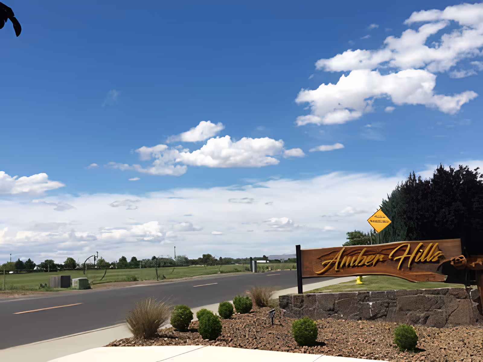Entrance sign reading "Amber Hills" by a roadside with landscaped shrubs and a blue sky with clouds.