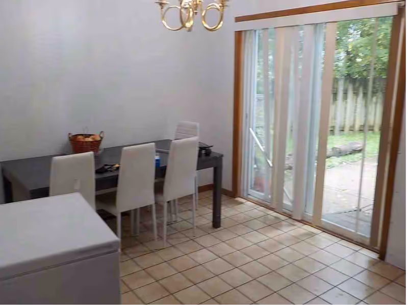 Dining area with a black rectangular table and four white chairs. A basket with fruit is on the table. There is a sliding glass door with vertical blinds leading to an outdoor patio. The floor is tiled and there is a chandelier hanging from the ceiling.