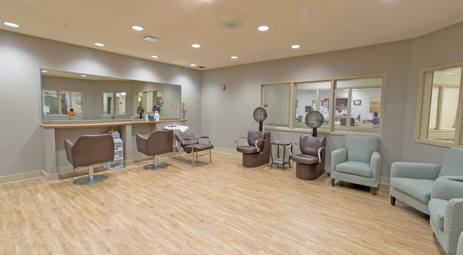 Interior view of a hair salon area in a senior living facility with brown salon chairs, hair dryers, a large mirror, and light blue armchairs. The room has wood flooring and beige walls with windows looking into an adjacent office area.
