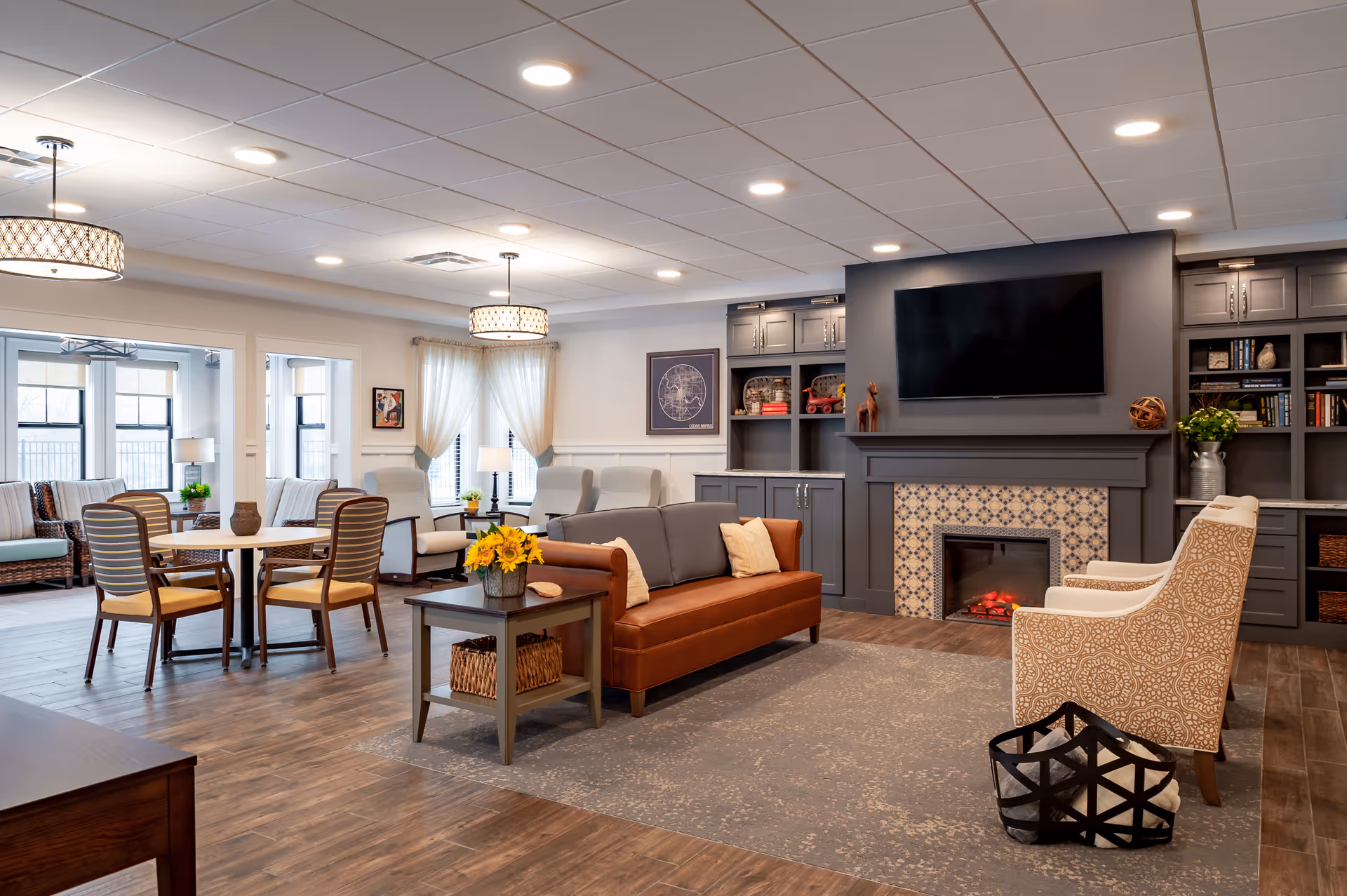 A spacious and well-lit living room area in a senior living facility featuring a brown leather sofa with pillows, two patterned armchairs, a wooden coffee table with a basket and a vase of sunflowers, a fireplace with decorative tiles, built-in shelves with books and decor, a large flat-screen TV mounted above the fireplace, and a dining area with a table and chairs near large windows with sheer curtains.