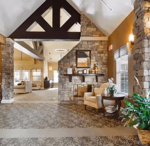 A cozy living room area with a stone fireplace, beige armchair, round wooden side table with a white orchid plant, and a large green potted plant. The room features high vaulted ceilings with dark wooden beams, warm lighting, and windows with blinds allowing natural light to enter. In the background, there is an open space with additional seating and large windows.