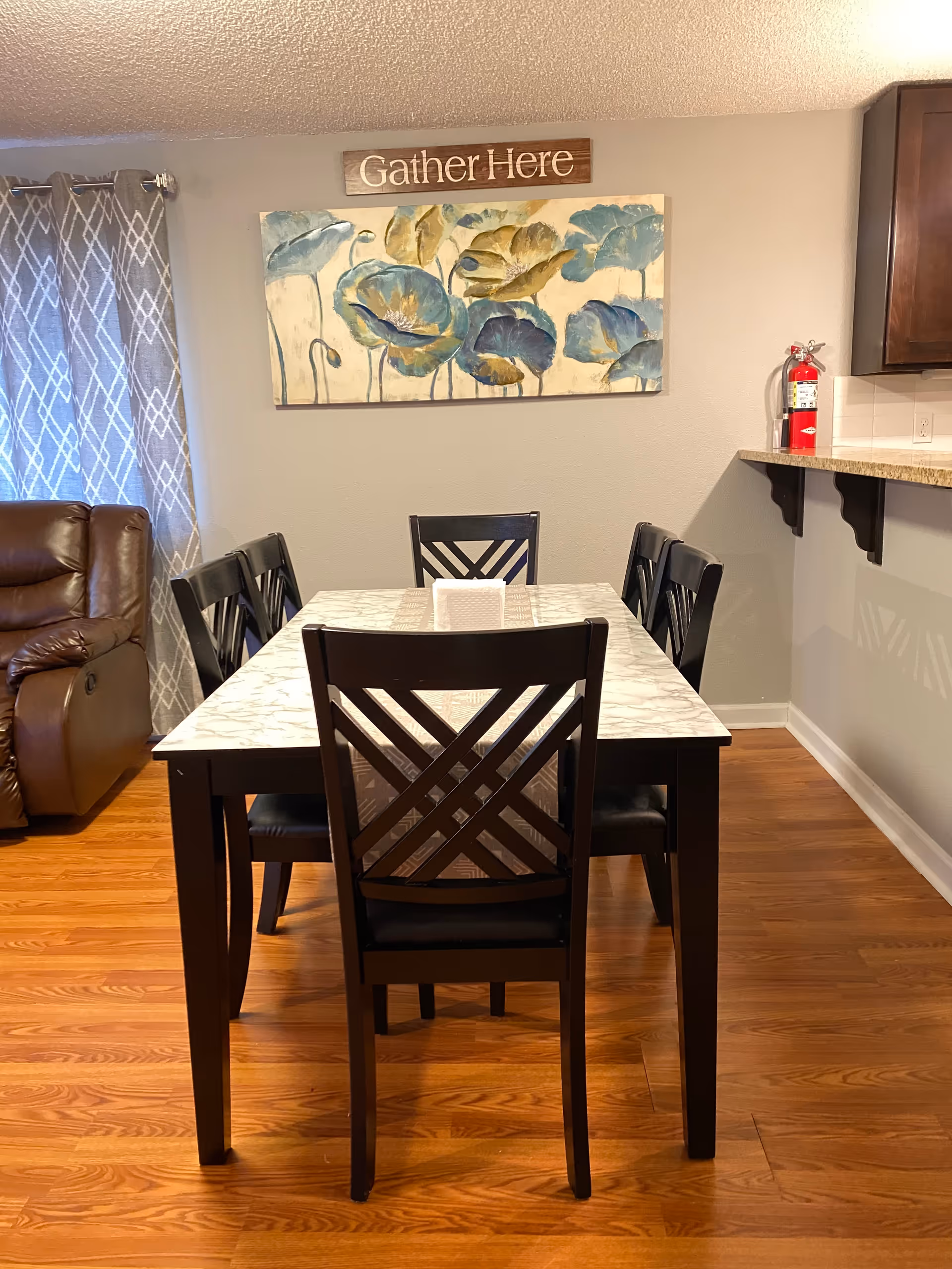 Dining area with a rectangular marble-top table surrounded by six dark chairs beneath a floral wall painting and a 'Gather Here' sign.