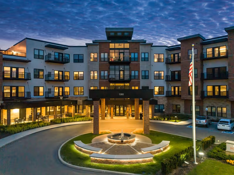 Exterior view of a modern senior living facility building at dusk with multiple windows lit, a circular driveway with a central fountain, an American flag on a flagpole, and some parked cars.