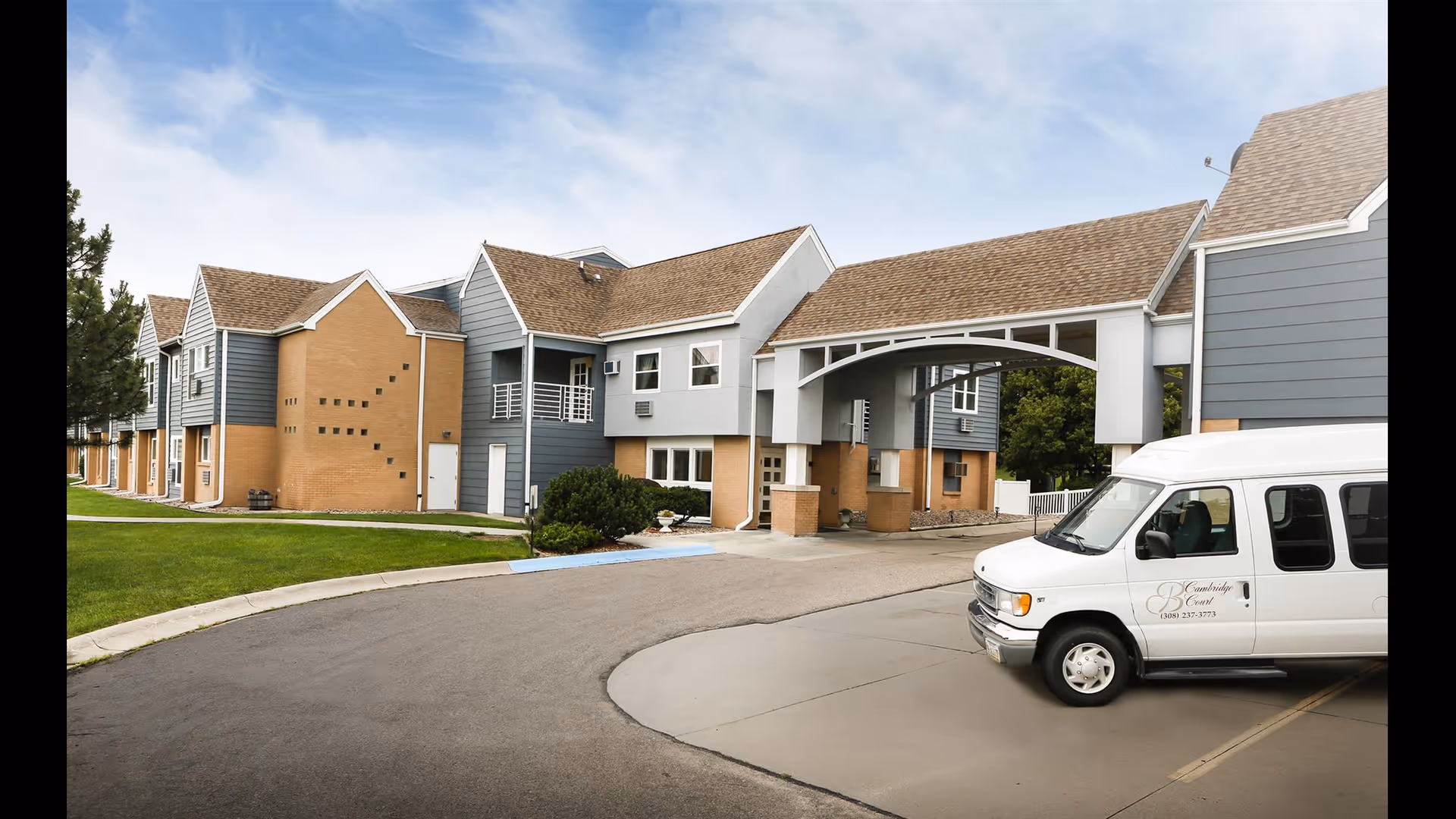Exterior view of Cambridge Court Assisted Living facility showing a multi-story building with blue and beige siding, a covered entrance, and a white van parked in front with the facility's name and phone number on the side.
