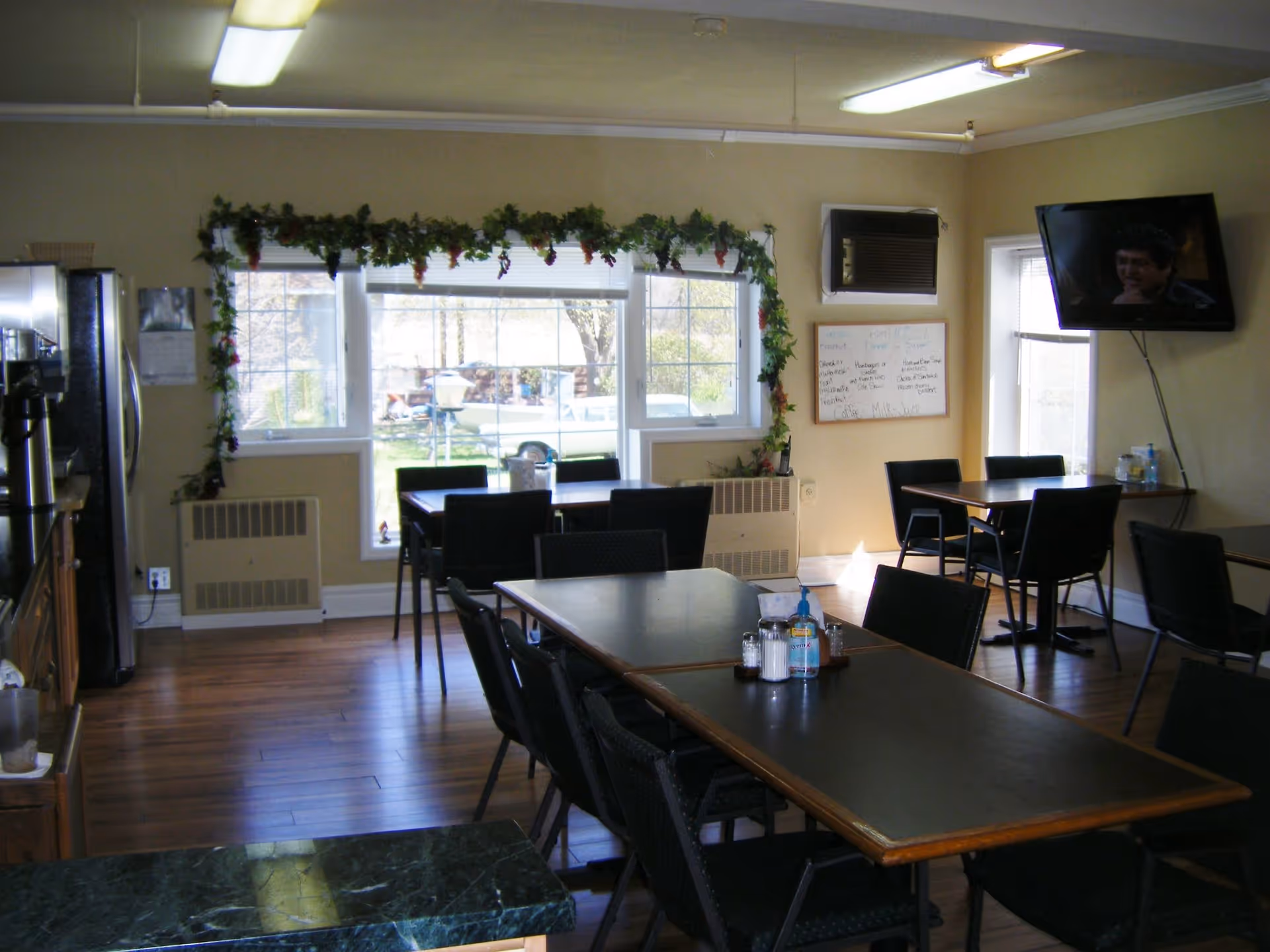 Interior view of a dining room with several black tables and chairs arranged neatly. There is a large window decorated with green garlands and red berries, allowing natural light to enter. A flat-screen TV is mounted on the wall to the right, and a whiteboard with writing is visible below it. The floor is wooden, and there is a refrigerator and coffee machine on the left side of the room.