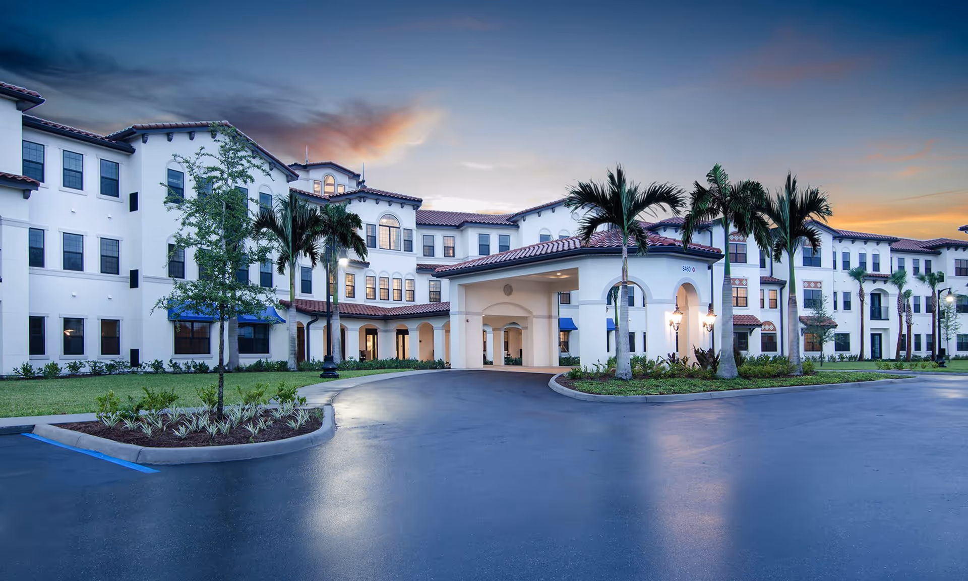 Exterior view of American House Coconut Point, a large multi-story senior living facility with white walls, red-tiled roofs, palm trees, and a covered entrance driveway at dusk.