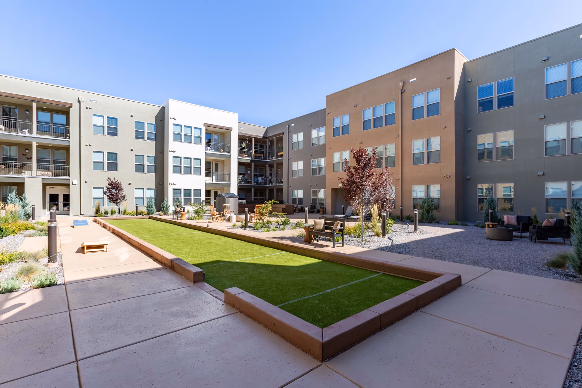 Outdoor courtyard area of a senior living facility with a bocce ball court in the center, surrounded by paved walkways, seating areas with chairs and benches, small trees, and multi-story residential buildings in the background under a clear blue sky.