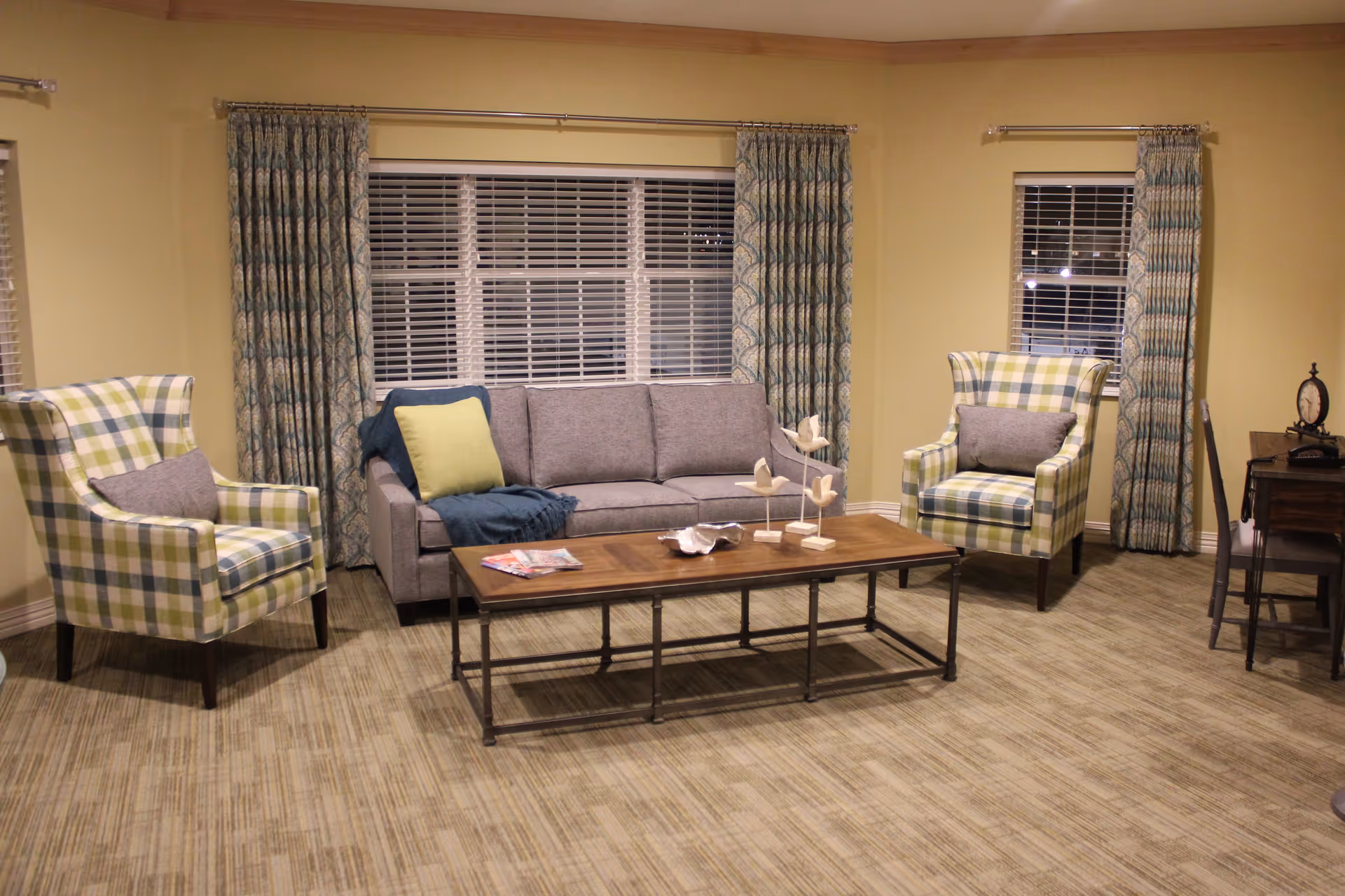 A cozy living room with a gray sofa adorned with a yellow pillow and a blue throw blanket, flanked by two plaid armchairs with gray cushions. A wooden coffee table with decorative items and magazines sits in front of the sofa. The room has large windows with patterned curtains and light-colored walls and flooring.