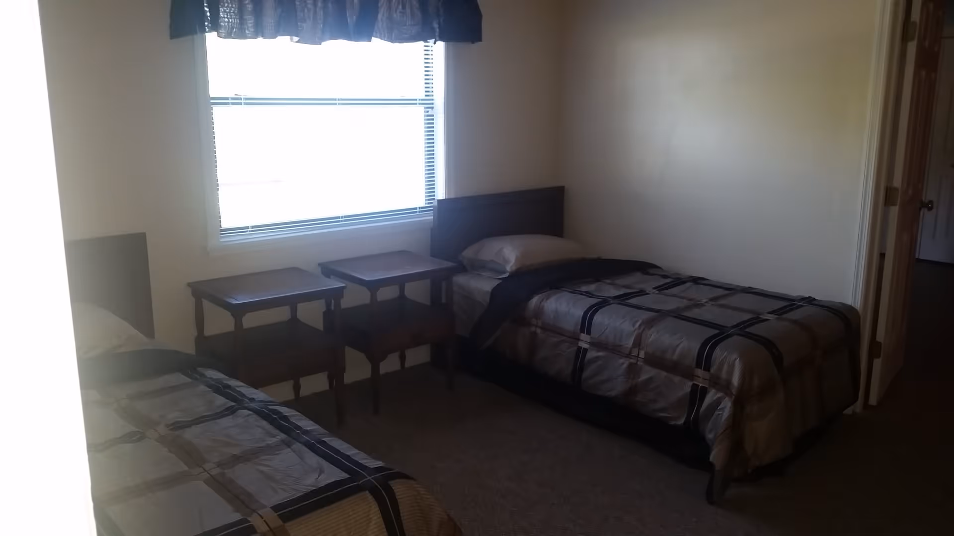 Simple bedroom with two twin beds, a window with blinds and two small wooden bedside tables.