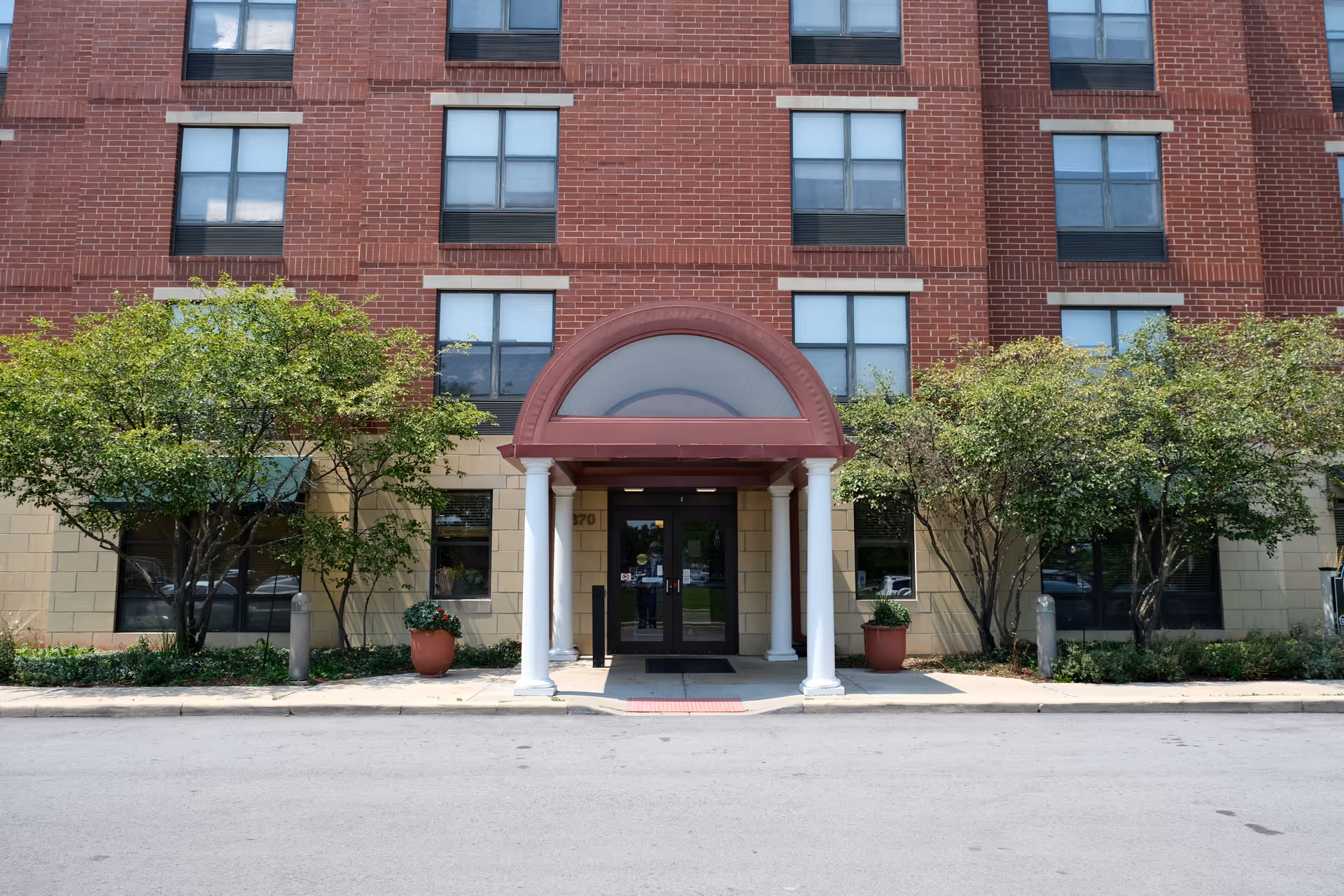 Front entrance of a brick building with a maroon arched canopy supported by white columns. There are green trees and potted plants on either side of the entrance, and multiple windows are visible on the building facade.