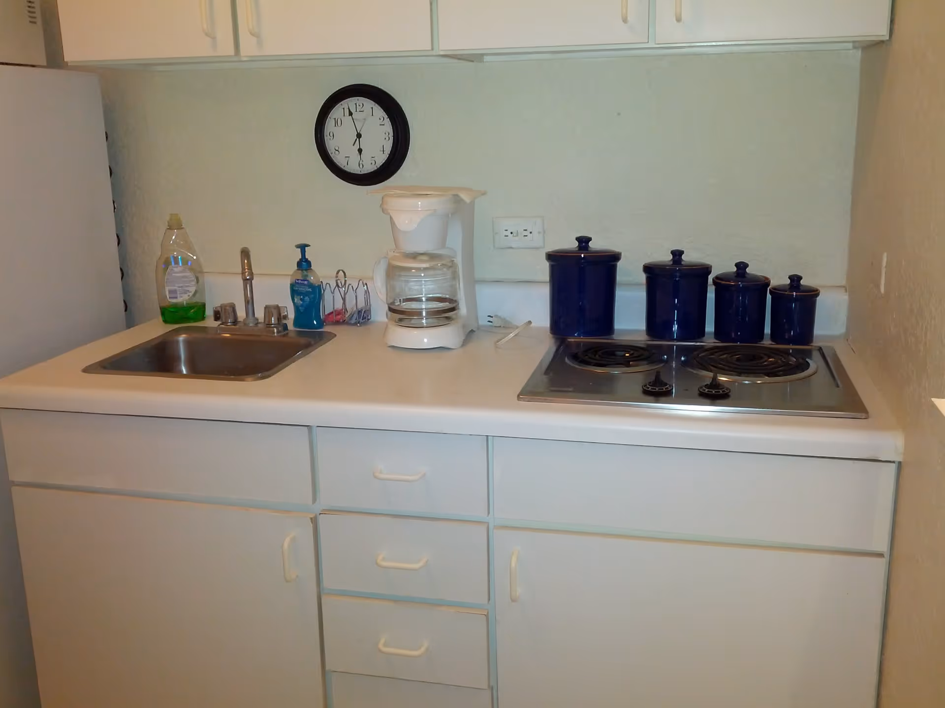 A small kitchen area with a white countertop featuring a stainless steel sink, a white coffee maker, four blue canisters, and a two-burner electric stove. Above the countertop are white cabinets, and a round black wall clock is mounted on the wall showing the time as 12:30. There is a bottle of dish soap and a hand soap dispenser near the sink.