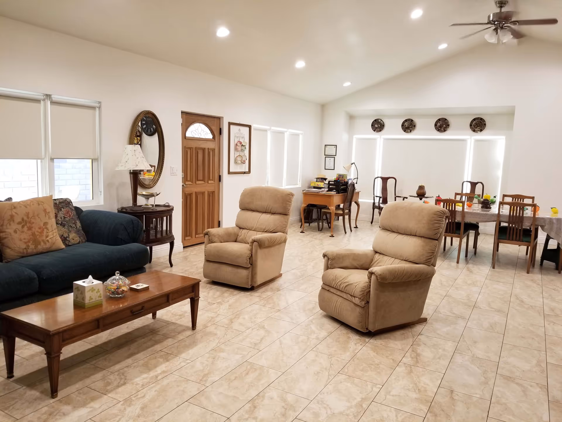 Bright open living room with two beige recliners, a green sofa, wooden coffee table and a dining area in the background on a tiled floor.