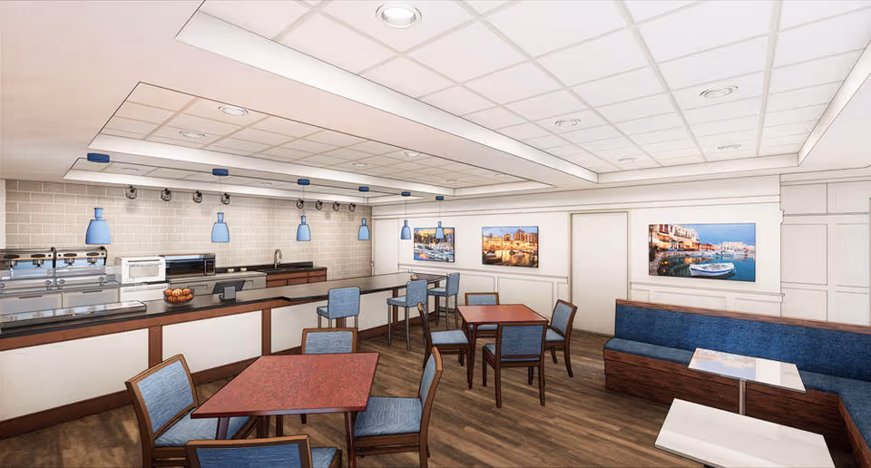 Interior view of a senior living facility dining area with several tables and chairs, a counter with bar stools, blue pendant lights hanging from the ceiling, and framed pictures of waterfront scenes on the walls.