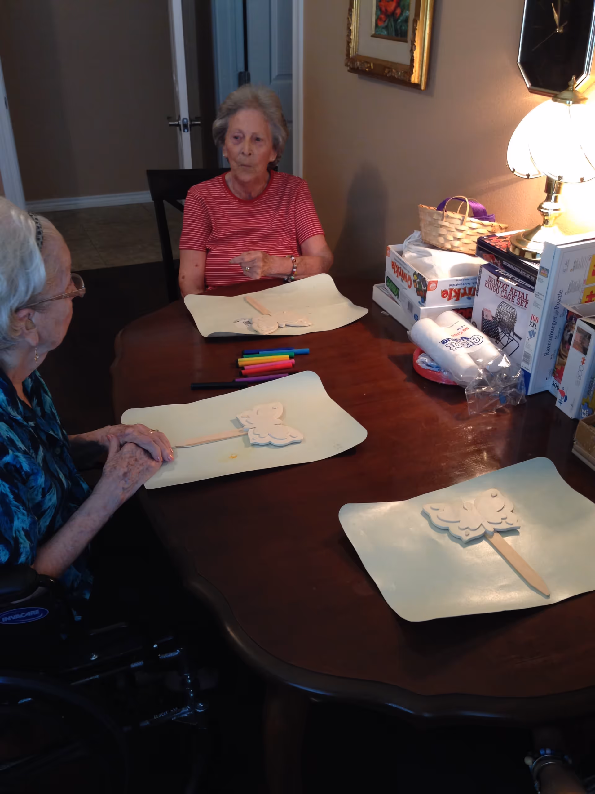 Two elderly women sitting at a wooden table engaged in a craft activity with butterfly-shaped wooden pieces and colored markers. One woman is in a wheelchair, and there are board games and a lamp on a side table in the background.
