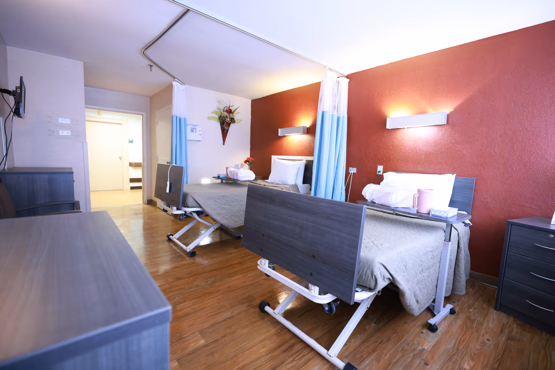 A rehabilitation center room with two hospital beds separated by a blue and white privacy curtain. Each bed has a beige blanket, white pillows, and an overbed table with items such as a pink pitcher and tissues. The room has wooden flooring, a red accent wall with two wall-mounted lights, a small dresser, and a wall-mounted TV.