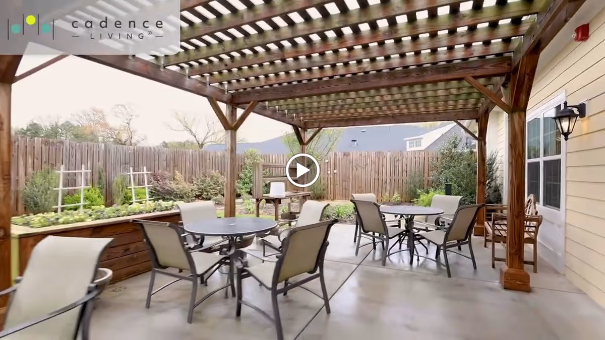 Outdoor patio area with a wooden pergola overhead, several round tables with chairs arranged underneath, a wooden fence surrounding the space, and garden beds with plants along the fence.