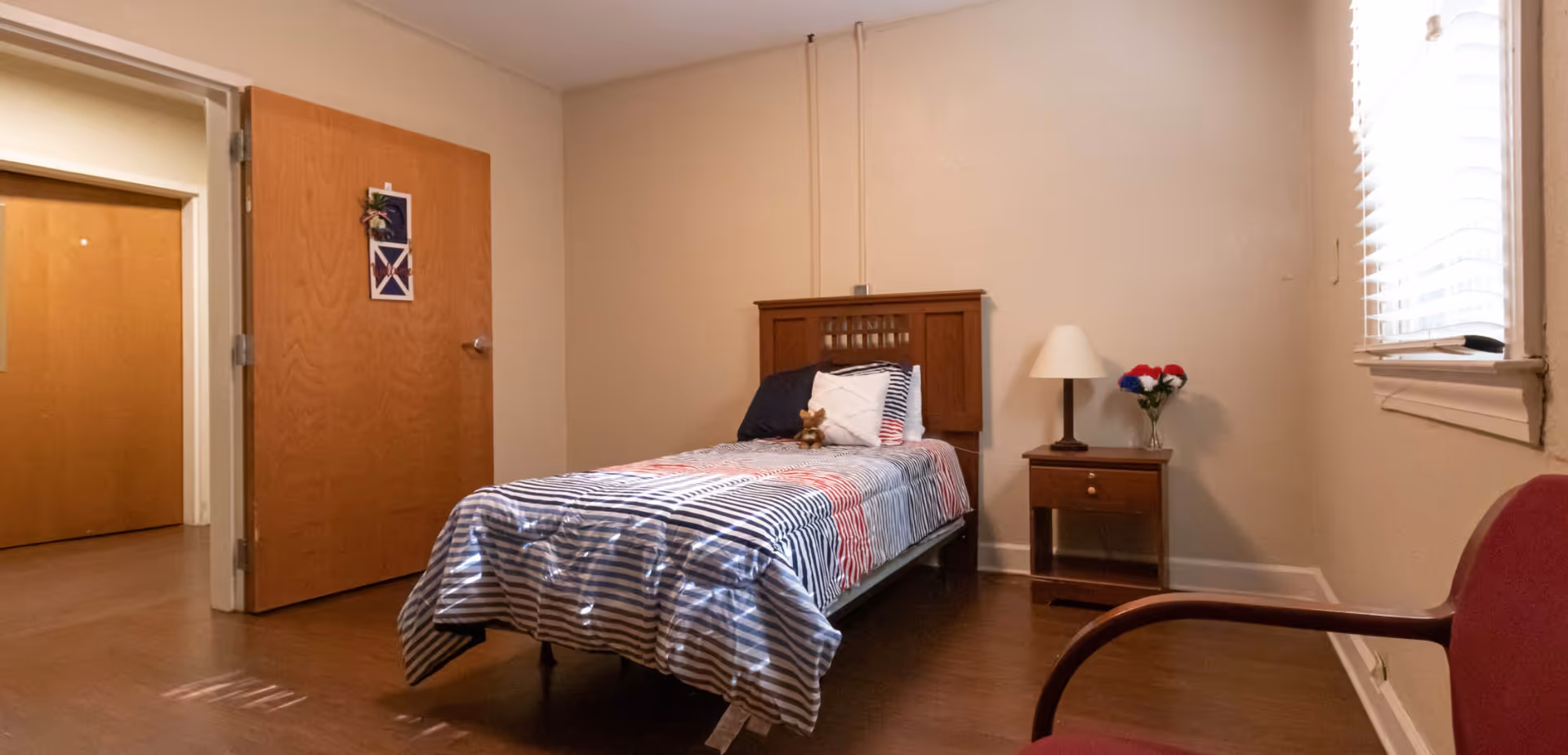 A simple single-occupancy bedroom with a twin bed dressed in striped bedding, a wooden headboard, nightstand with a lamp and flowers, a chair, and an open door.