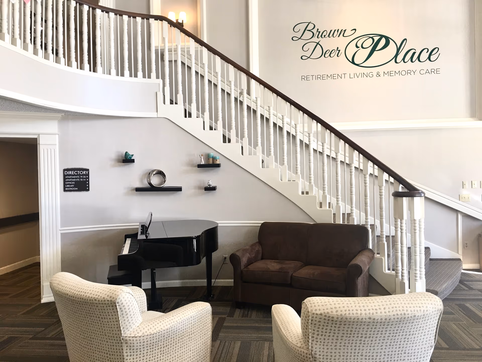 Interior view of a senior living facility lobby area with a curved staircase, a black grand piano, a brown loveseat, and two white patterned armchairs. The wall features the text 'Brown Deer Place Retirement Living & Memory Care' and a small directory sign. Decorative shelves with small items are mounted on the wall above the piano.